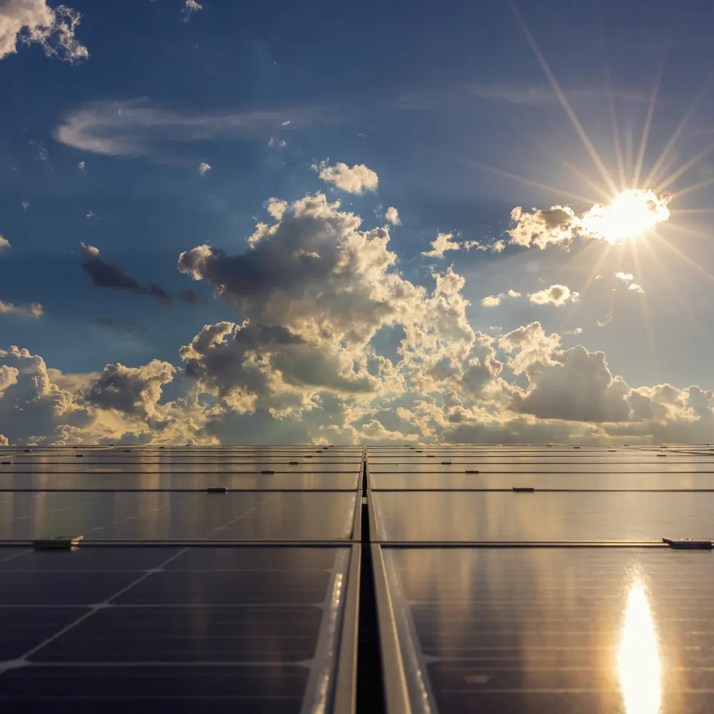 A view with clouds in the sky along with the sun and solar panels on the ground.